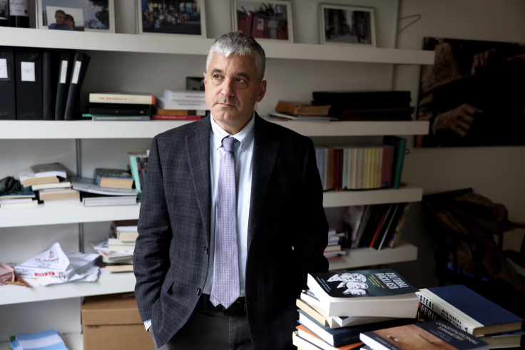 Man in suit stands in office with hands in his pockets.