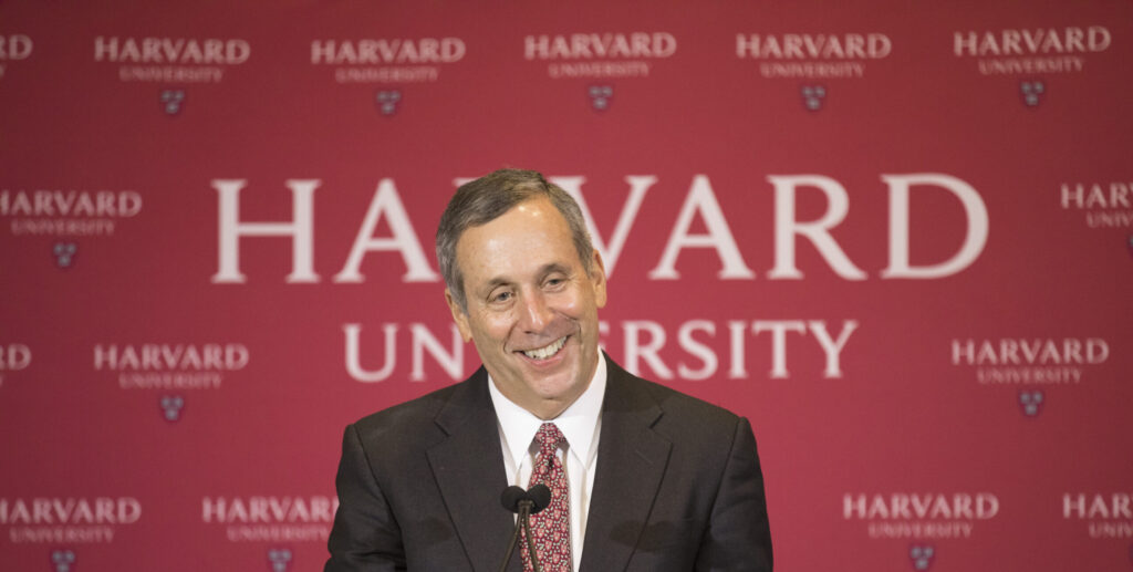 Man in a suit in front of a Harvard University banner.