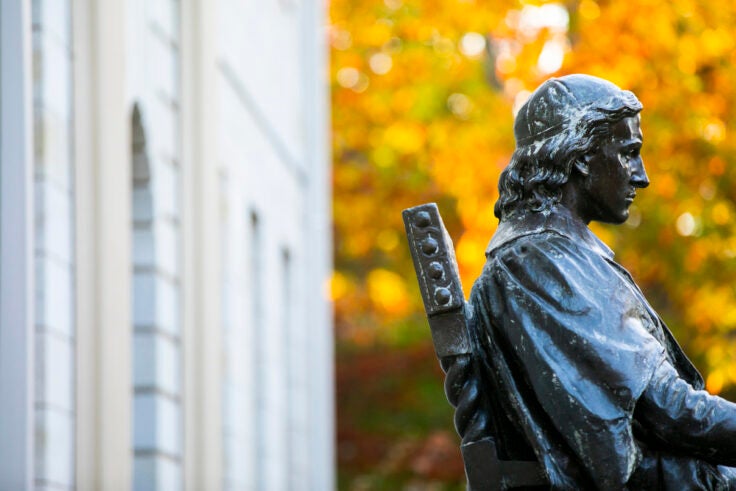 John Harvard statue in front of yellow fall leaves.