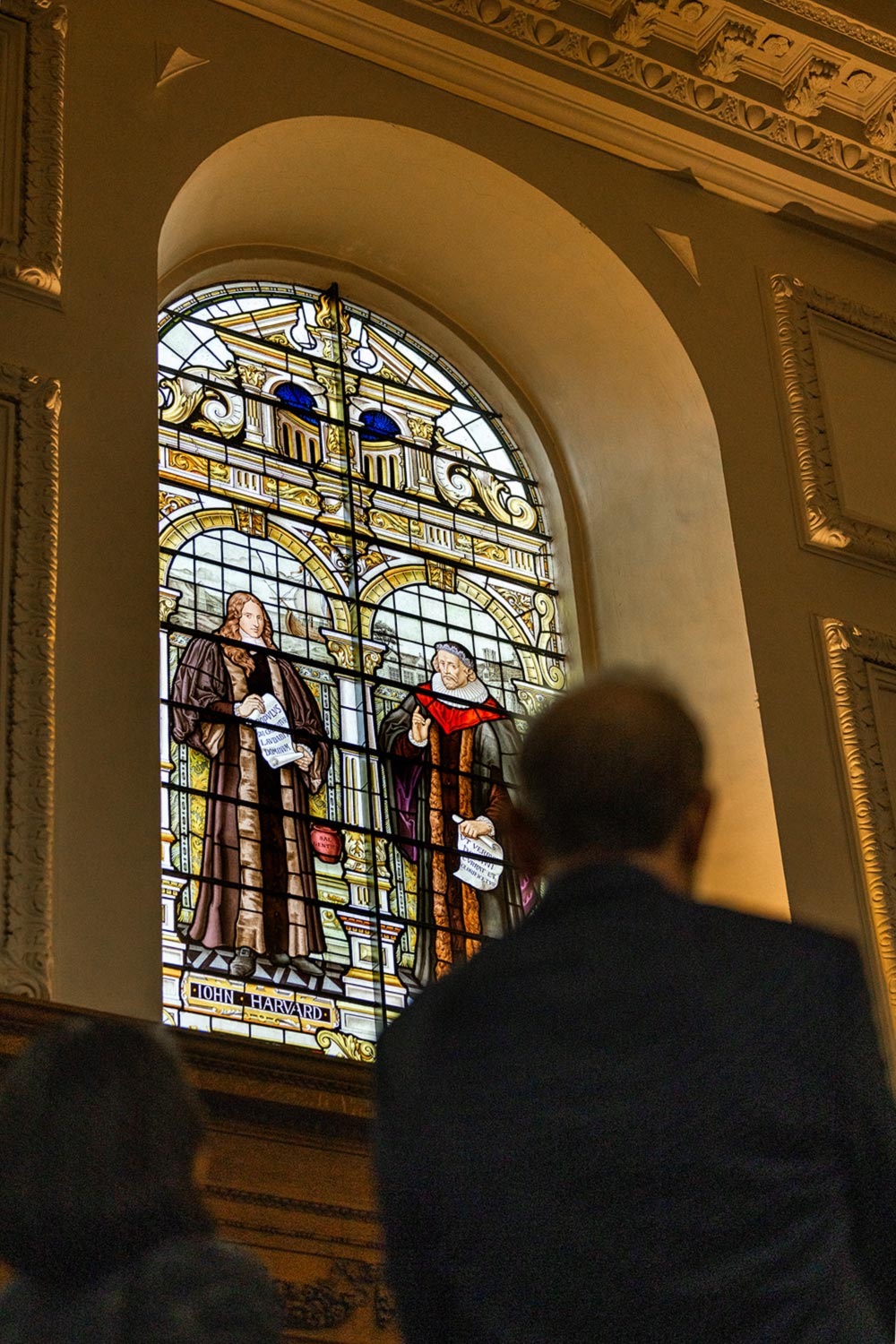 A man looks up at a stained glass window depicting John Harvard and another scholar.