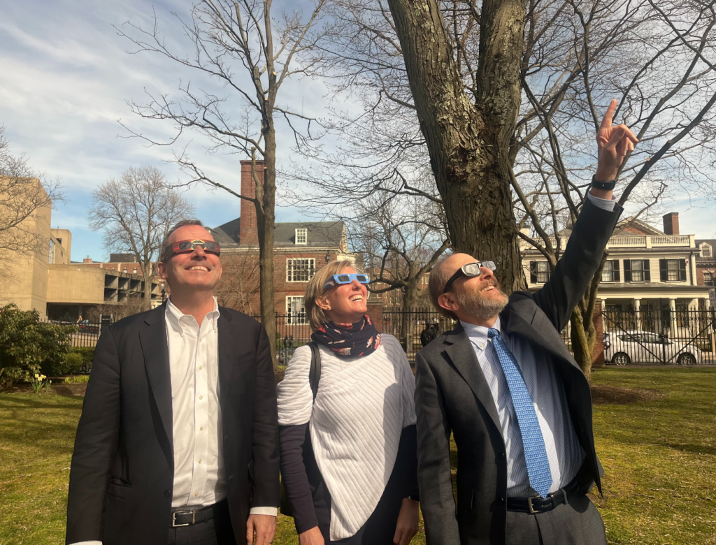 Three people wearing solar glasses look skyward at the total eclipse. 