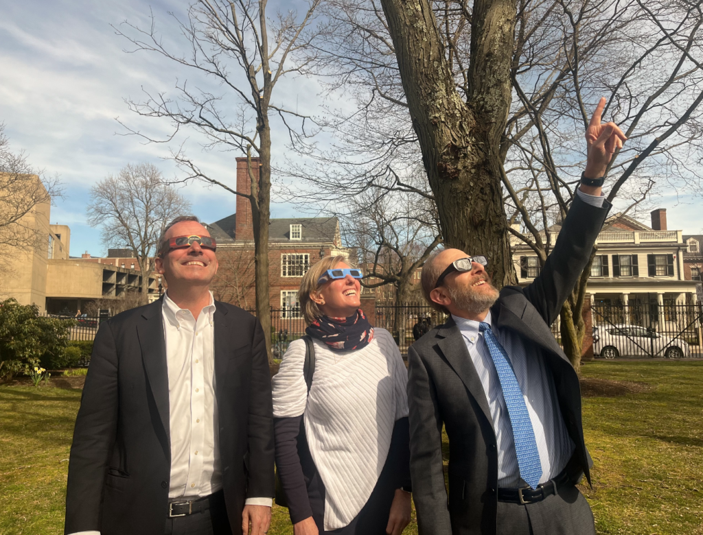 Three people wearing solar glasses look skyward at the total eclipse.