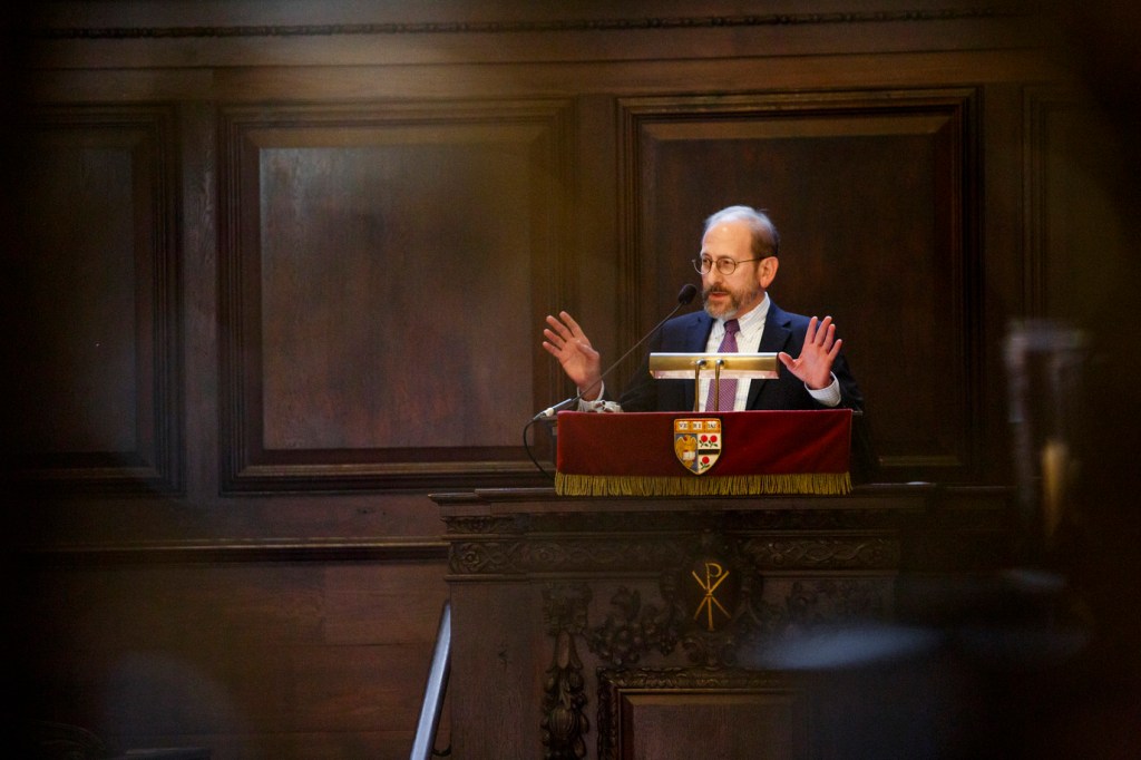 Alan Garber speaking at a podium raising both of his hands.