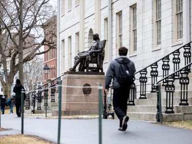 Young man walks in front of the John Harvard statue.