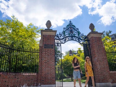 Two student walk through an open gate, with green summer leaves on trees behind them