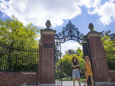 Two student walk through an open gate, with green summer leaves on trees behind them