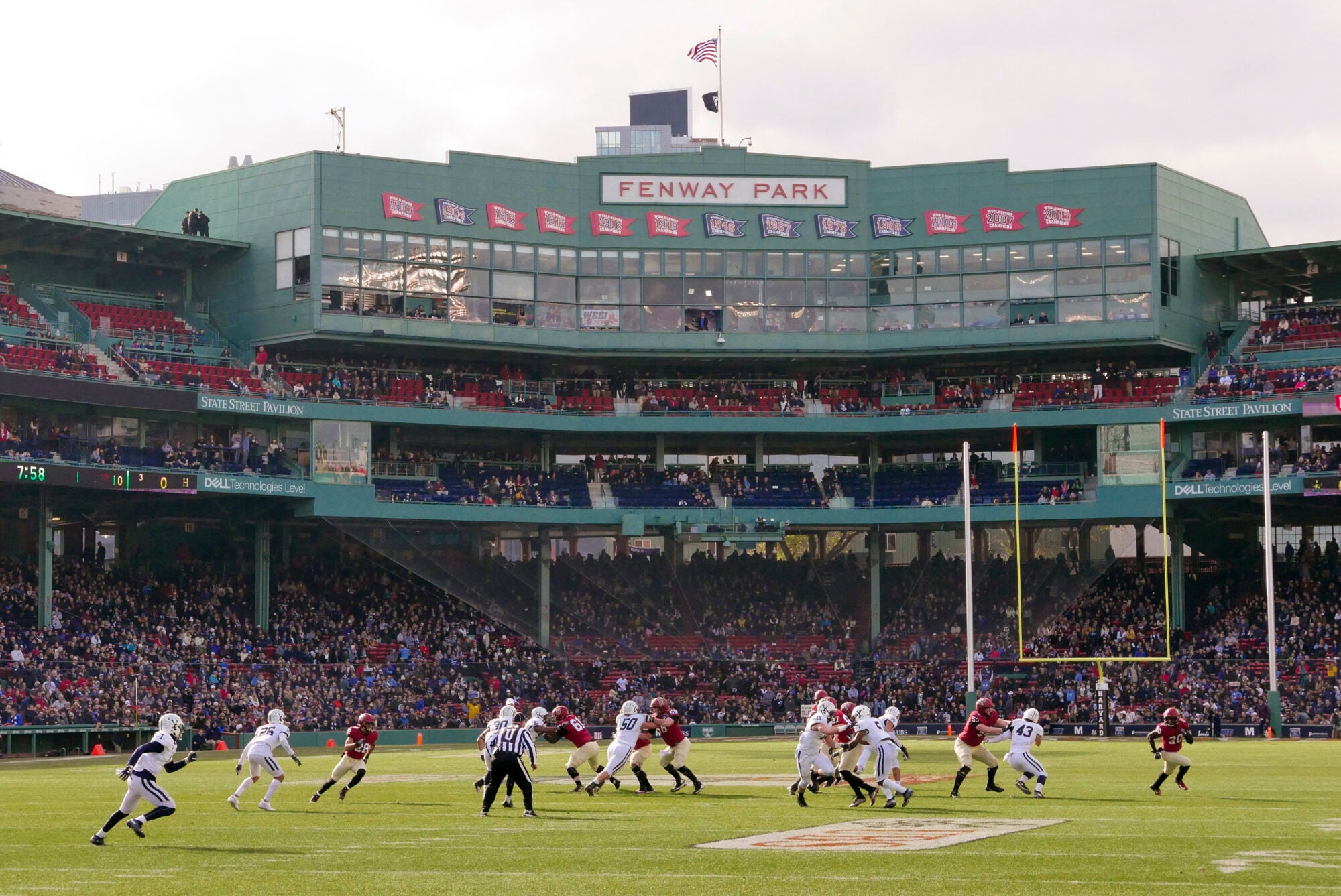 Harvard and Yale football teams playing in front of the large, green Fenway Park sign in the stadium.