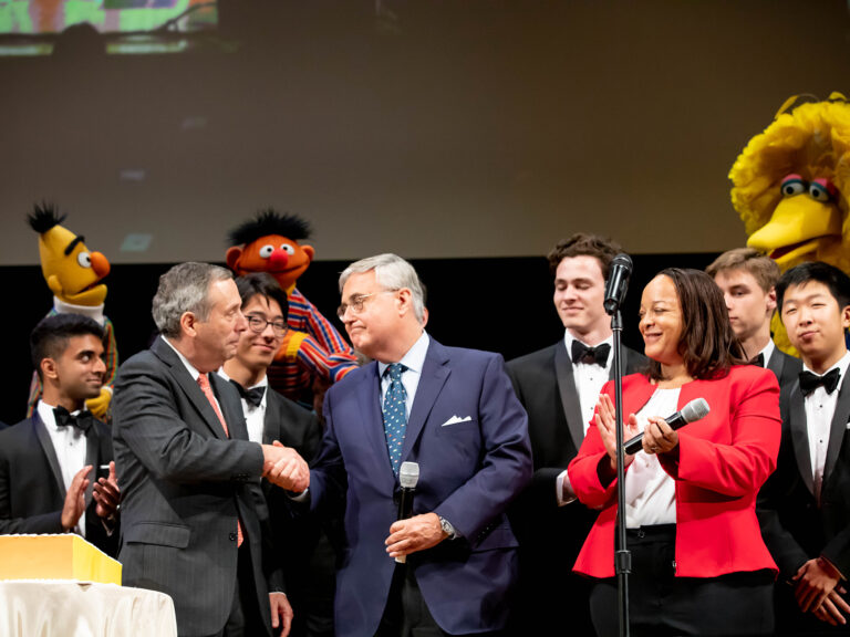 Two men on stage shaping hand with people and Muppets surrounding them.