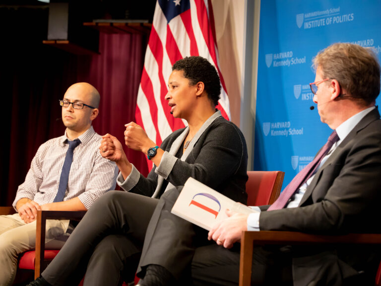 Three people seated on a stage talking to one another in front of an American flag.