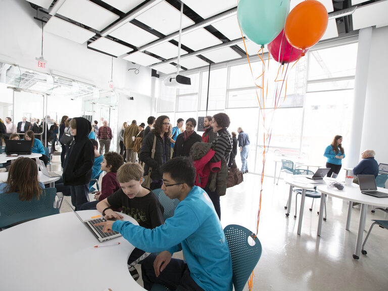 Two people sit at a table with a laptop, behind them is a group in a large sunny room with balloons.