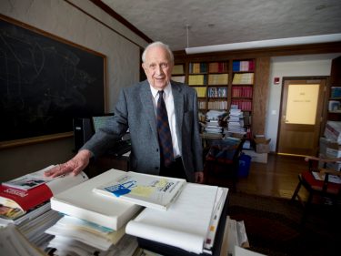 An older man standing in his office behind a pile of books and papers.