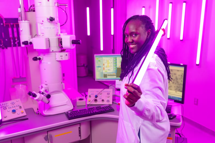 A woman wearing a white lab coat stands in a laboratory holding a fluorescent tube. The room is lit in a pink/purple color.