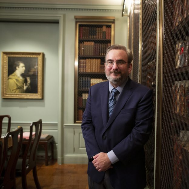 A man stands in a library, with books and a painting behind him