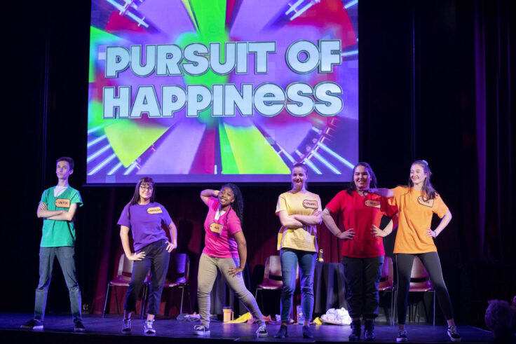 Six teenagers on stage with a large sign that reads "Pursuit of Happiness" behind them