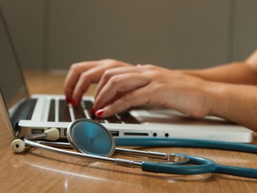 Someone typing on a laptop with a stethoscope next to it