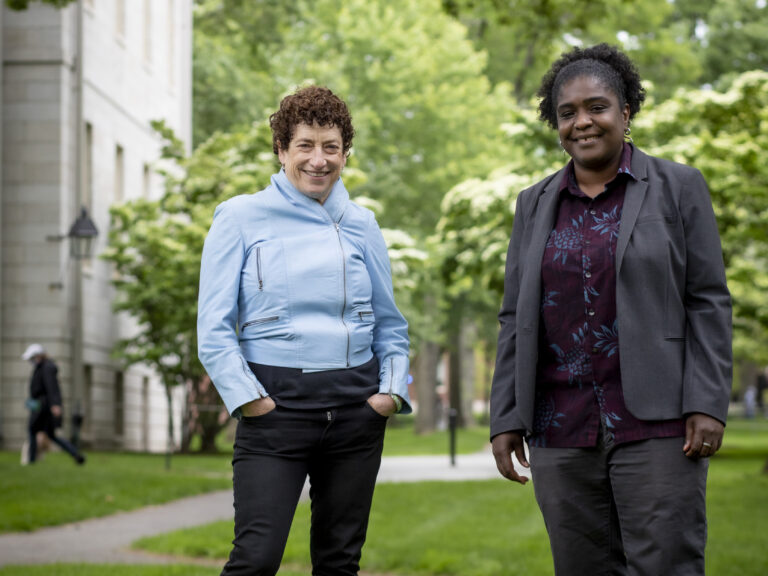 Two women stand together on green grass with green trees behind them