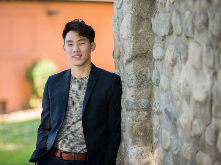 A student wearing a blazer leans against a tree trunk