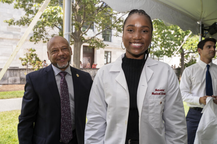 A student wearing her white coat smiles, while a faculty member looks on