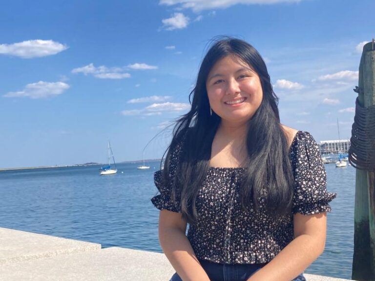A student sits in front of the water, with a blue sky and puffy white clouds in the sky