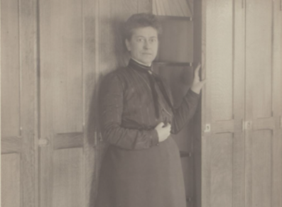 old image of woman standing by a bookshelf in turn of century dress.