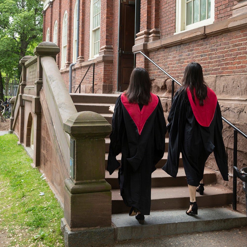 Two graduates wearing robes walk up steps to a brick building