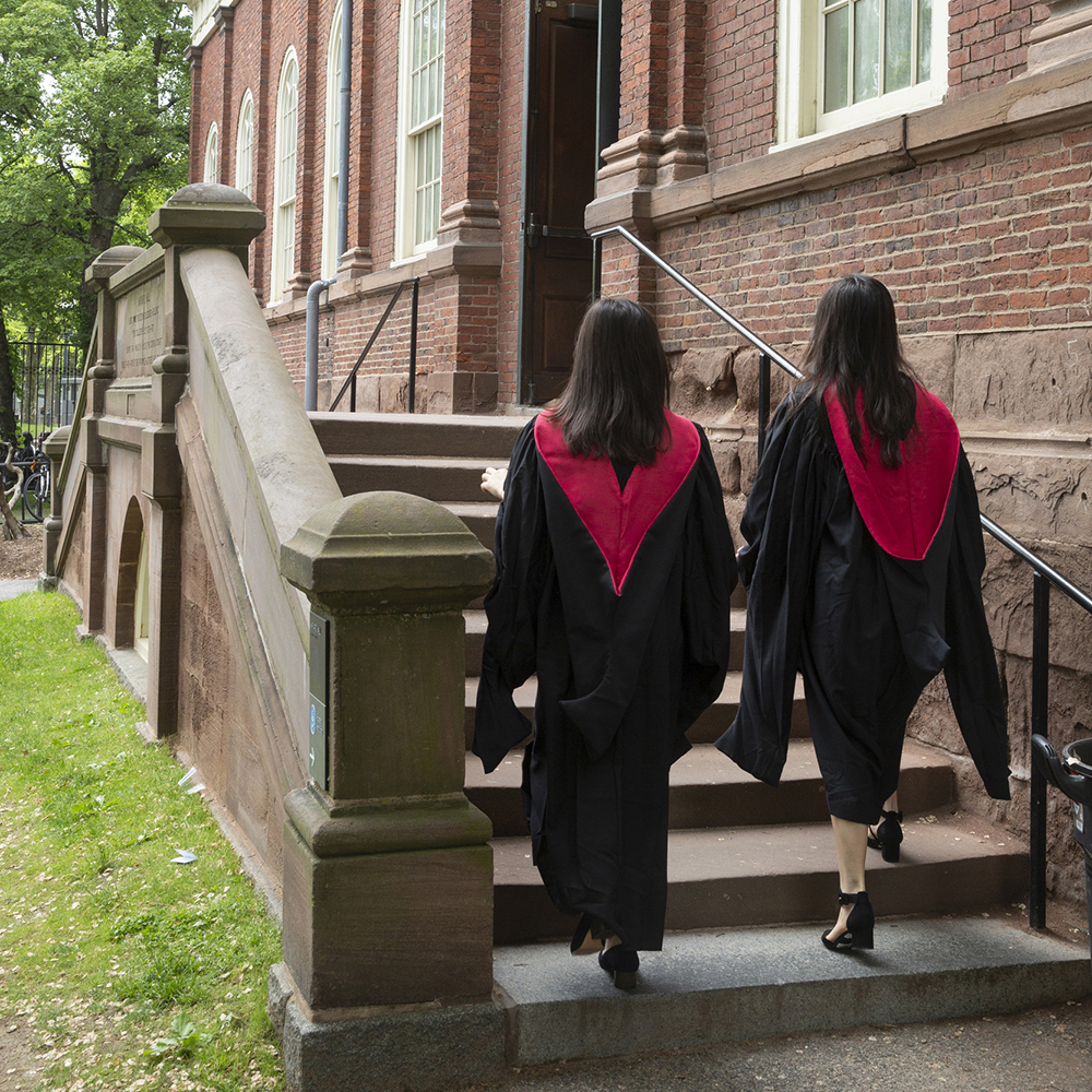 Two graduates wearing robes walk up steps to a brick building