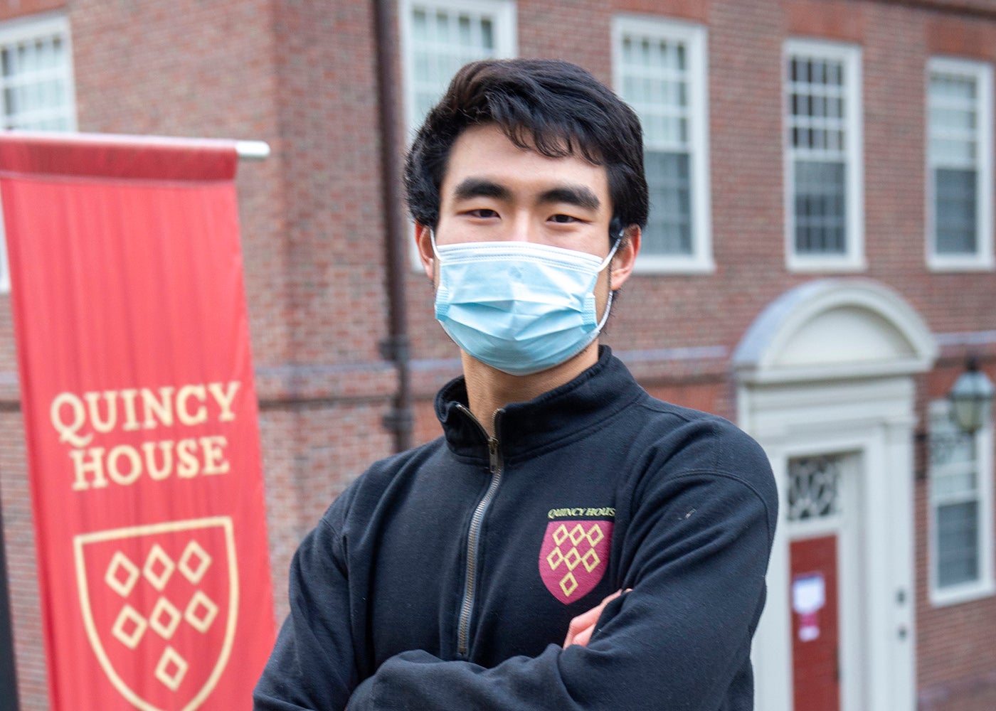 A student wearing a mask poses in front of a red Quincy House banner on campus