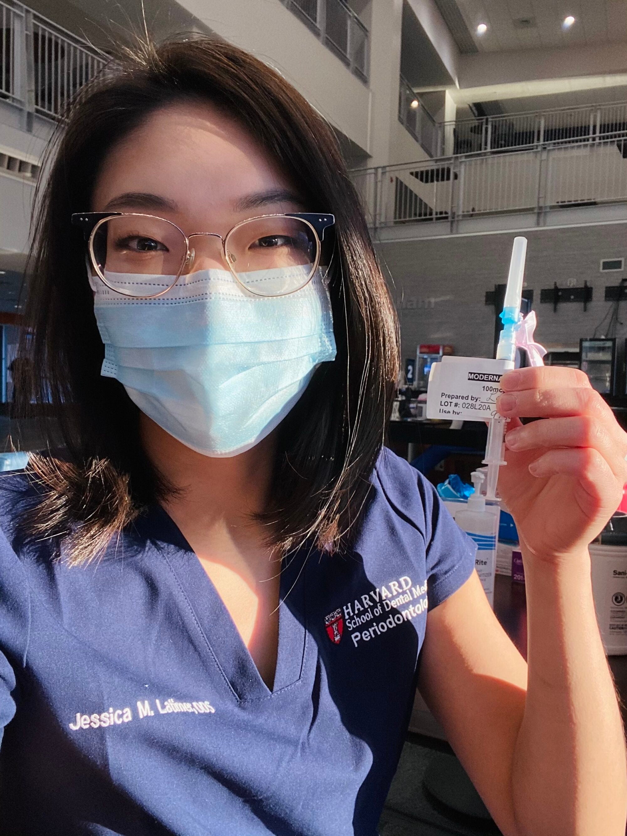 A young woman in blue scrubs holds a COVID-19 vaccine syringe