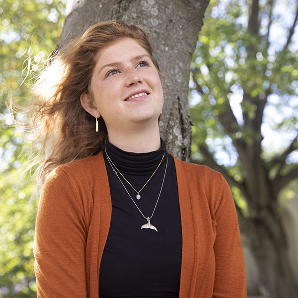 A woman stands in front of a tree with fall leaves, looking off into the distance