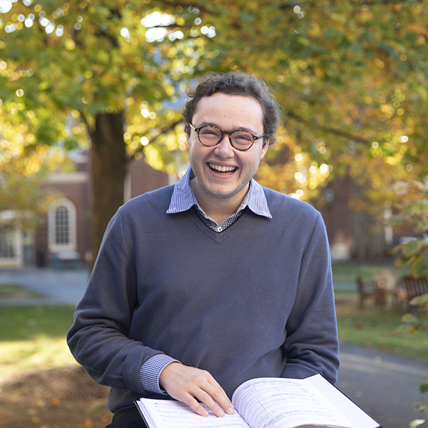 A student standing outside in front of fall foliage holds an open book