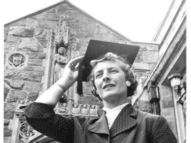 A black and white photo of a woman holding a graduation cap on her head outside a building with a cross on it