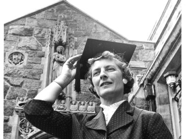 A black and white photo of a woman holding a graduation cap on her head outside a building with a cross on it