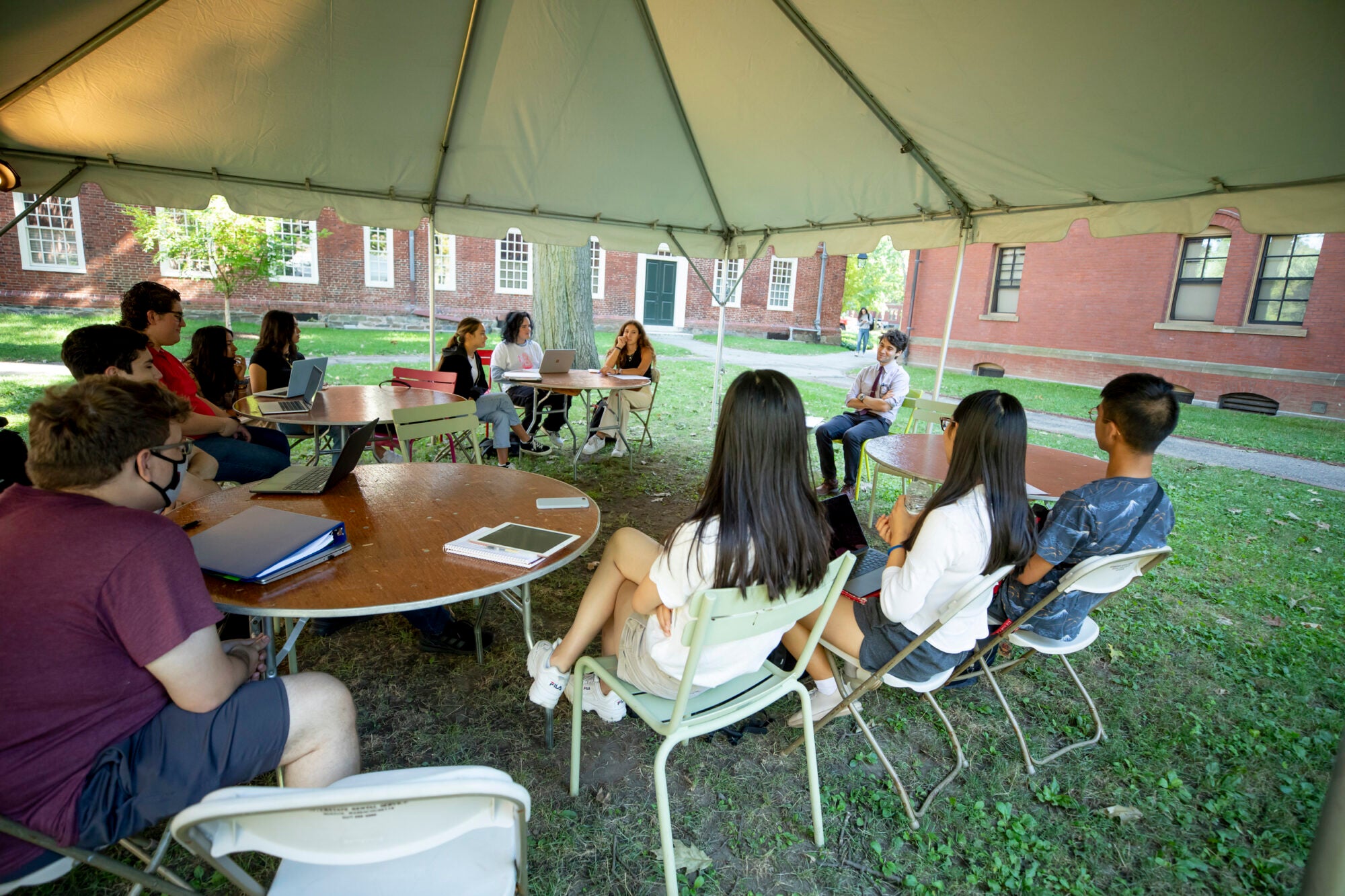 Students sit in chairs under a tent outside during class