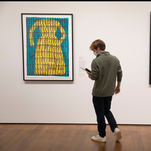 A student looks down at notes while studying artwork on the wall of a gallery