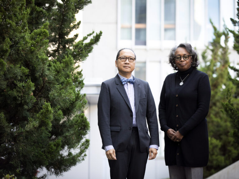 Two people stand together outdoors amongst trees with a white building behind them