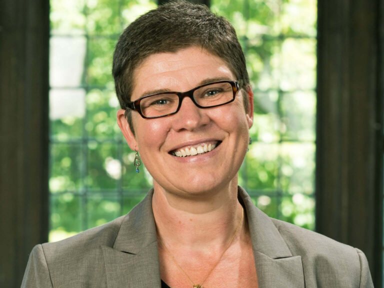 A woman wearing glasses and a blazer poses for a headshot in front of a window