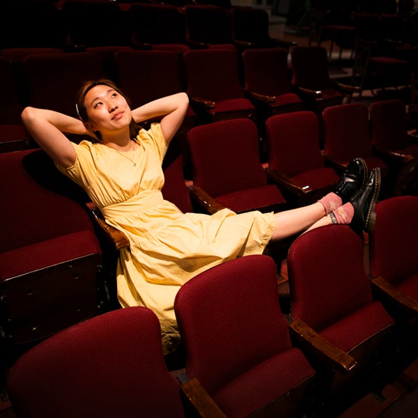 A student reclines in a theater seat with her hands behind her head and he feet up on the chair in front of her