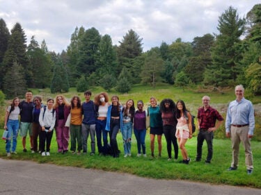 A group of students pose outdoors with their professor with trees on the horizon