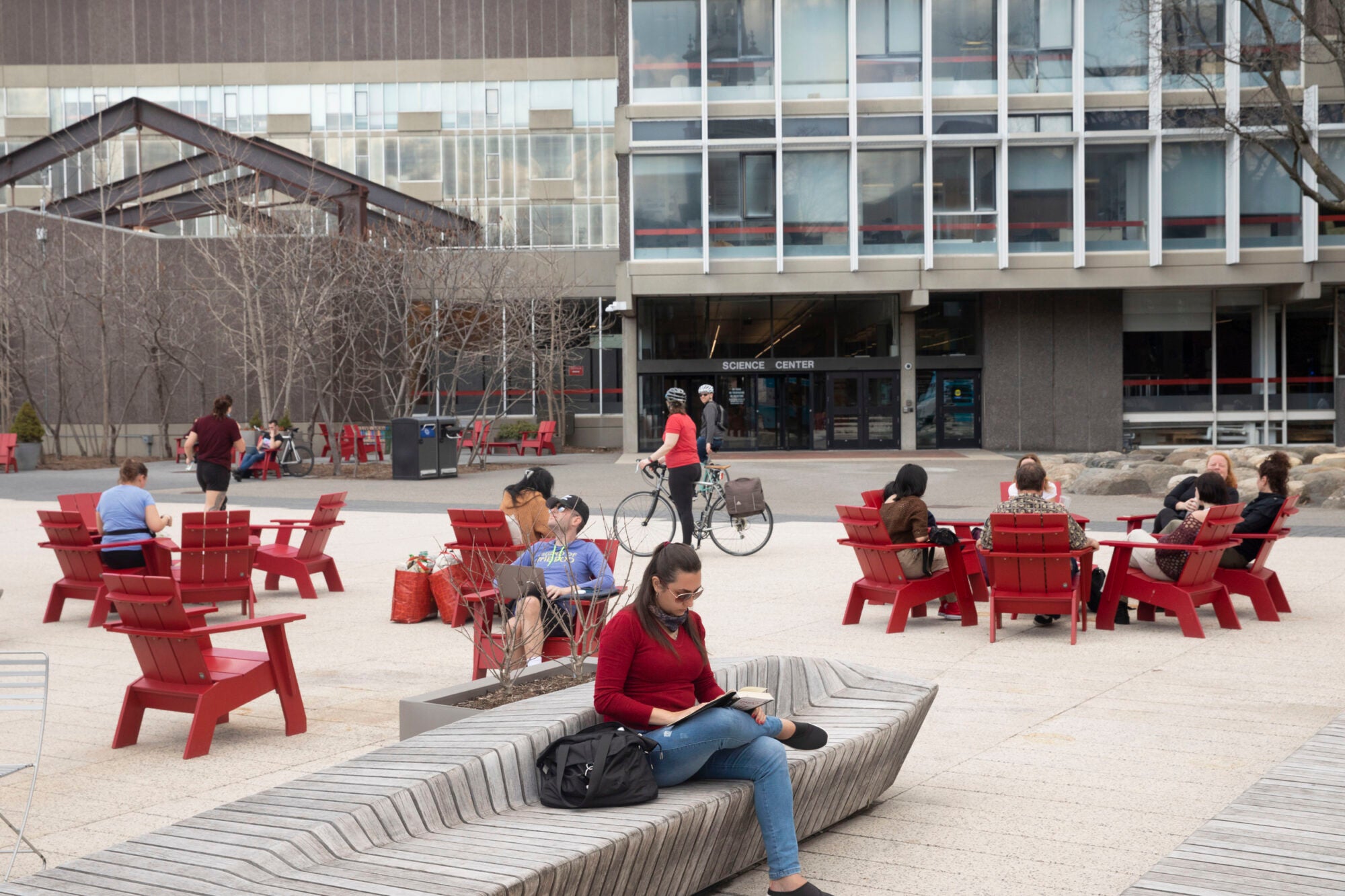 People sit on benches and in chairs on a plaza with a building behind them
