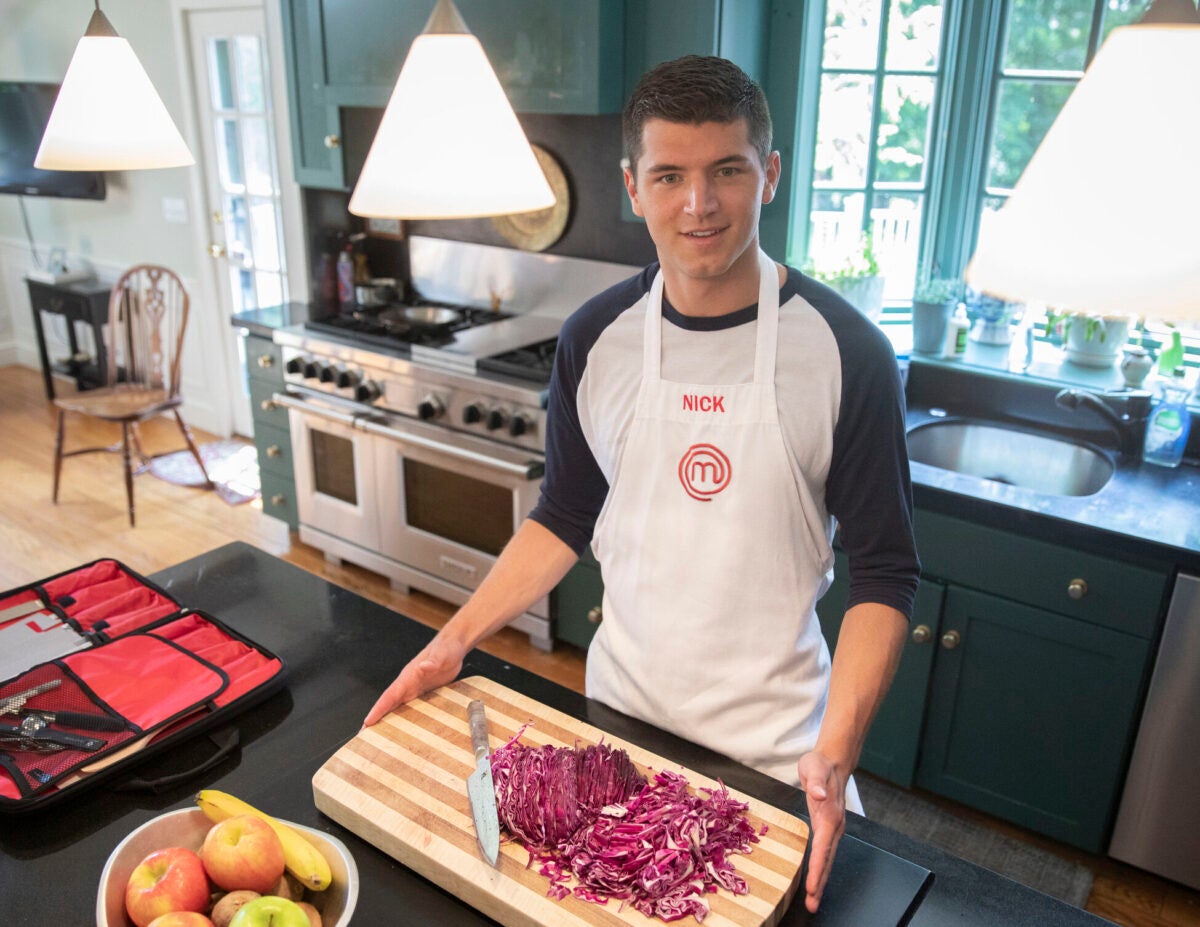 A student wearing an apron stands in a kitchen with a cutting board of purple cabbage in front of him