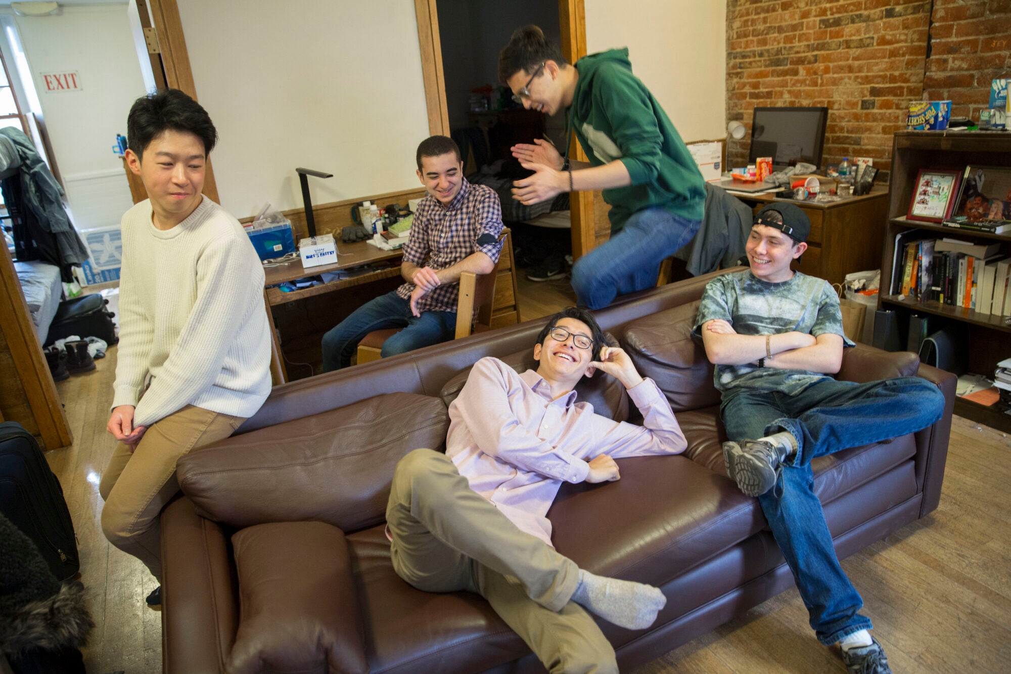 Five young men lounge and laugh on a couch and at a desk in a dorm room