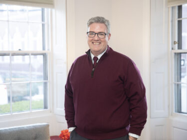 A man wearing a maroon sweater stands between two windows in an office