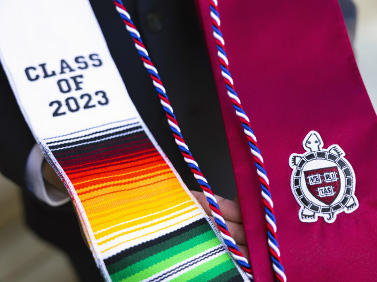 A student displays their graduation regalia, one side is crimson and one side is multi-colored with the words "Class of 2023" on it.