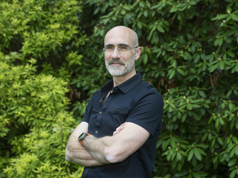 A man stands with his arms crossed in front of greenery