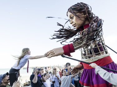 A large puppet and a young girl reach out to each other, touching hands.