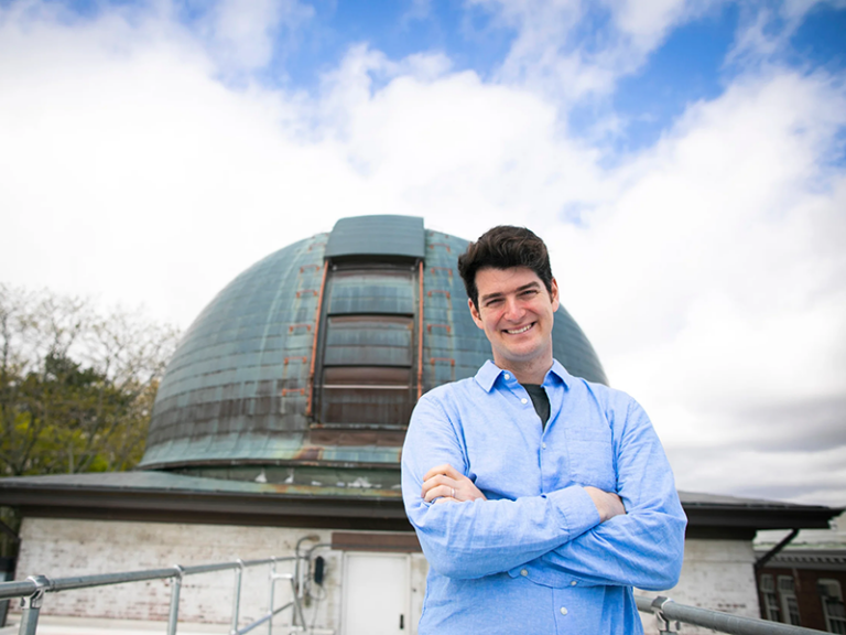 A young man stands in front of a planetarium with blue sky and clouds behind him