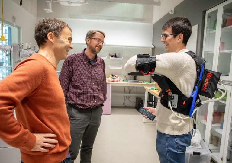 A man wears a robotic vest while two researchers look on