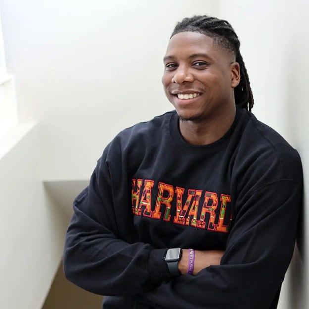 A man wearing a Harvard sweatshirt leans against a wall