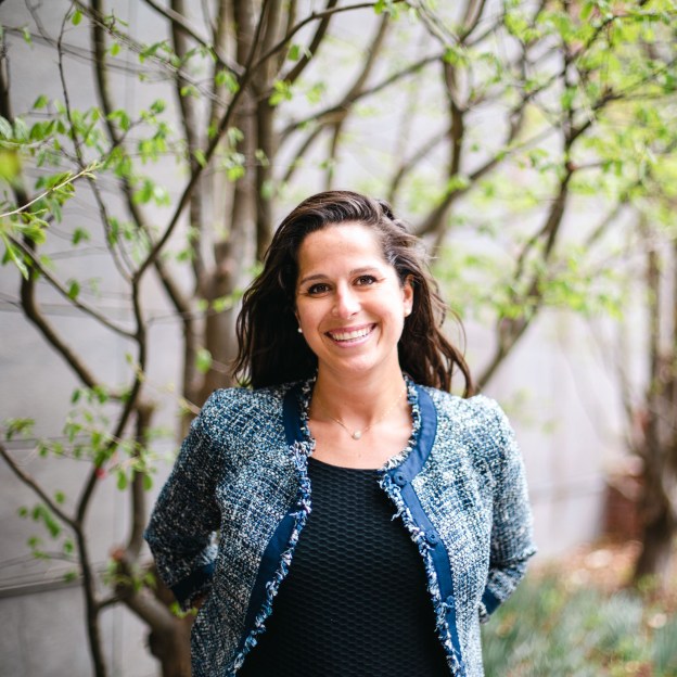 A smiling woman stands outside in front of a budding tree.