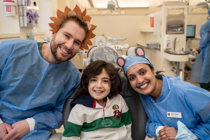 Two dental students wearing kid-friendly head bands pose with a child in a dental chair.